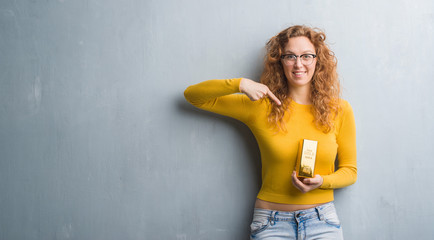 Young redhead woman over grey grunge wall holding gold ingot with surprise face pointing finger to himself