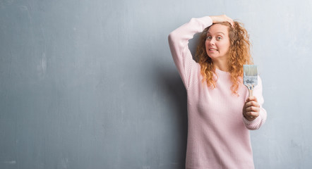 Young redhead woman over grey grunge wall holding paint brush stressed with hand on head, shocked with shame and surprise face, angry and frustrated. Fear and upset for mistake.