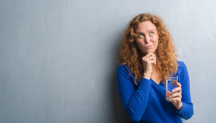 Young redhead woman over grey grunge wall drinking water serious face thinking about question, very confused idea - obrazy, fototapety, plakaty