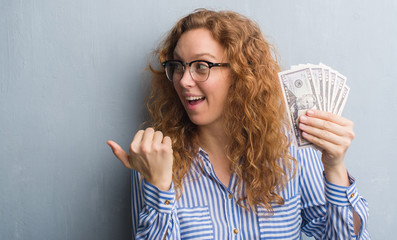 Young redhead woman over grey grunge wall holding dollars pointing and showing with thumb up to the...