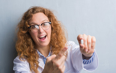 Young redhead doctor woman over grey grunge wall holding a pill very happy pointing with hand and...