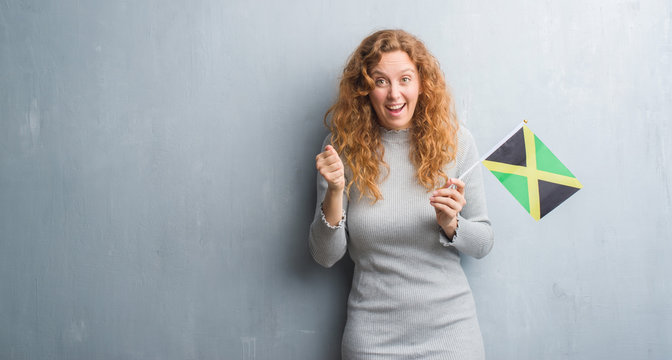 Young redhead woman over grey grunge wall holding flag of Jamaica screaming proud and celebrating victory and success very excited, cheering emotion