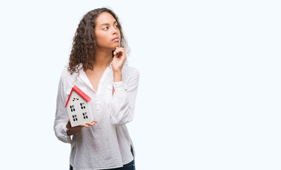 Young hispanic real state agent woman holding small house serious face thinking about question,...