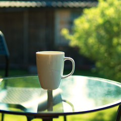 Cup of coffee on glass table with backdrop of sun light, grenn leafs, trees and house. Summer morning, selective focus.