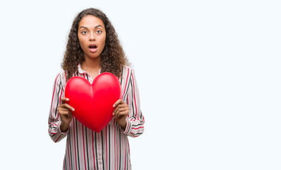 Young hispanic woman in love holding red heart scared in shock with a surprise face, afraid and excited with fear expression