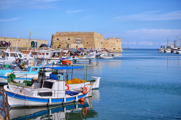 Heraklion, Crete Island - Greece. Traditional colorful fishing boats are docked at the old port of Heraklion city in front of the fortress Koules (castello a mare). Sunny day, blue cloudy sky