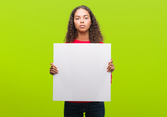 Young hispanic woman holding blank banner with a confident expression on smart face thinking serious