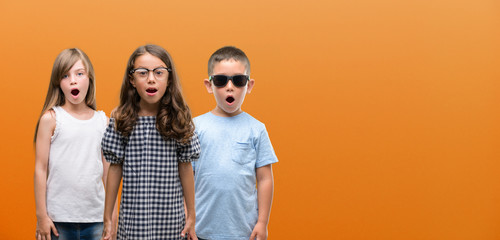 Group of boy and girls kids over orange background scared in shock with a surprise face, afraid and...