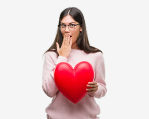 Young hispanic woman holding heart in love cover mouth with hand shocked with shame for mistake, expression of fear, scared in silence, secret concept