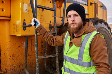 Waist up portrait of bearded worker posing with heavy yellow truck and  looking at camera, copy...