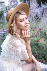 Young blonde is walking in a flower garden. A young woman is holding flowers and a straw hat. Purple. Lavender. Roses.