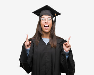 Young hispanic woman wearing graduated cap and uniform amazed and surprised looking up and pointing...
