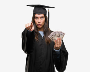 Young hispanic woman wearing graduated uniform holding dollars pointing with finger to the camera and to you, hand sign, positive and confident gesture from the front
