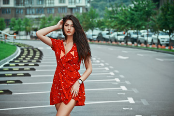 Young brunette girl standing and poses in red short dressn the city street, walking on empty parking place