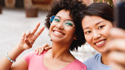 Two happy friends looking at smartphone and taking selfie, sitting in outdoor cafe