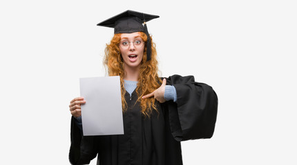Young redhead woman wearing graduate uniform holding degree with surprise face pointing finger to himself