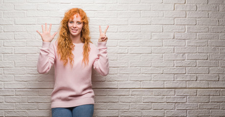 Young redhead woman standing over brick wall showing and pointing up with fingers number seven while smiling confident and happy.