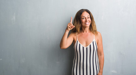 Fototapeta premium Middle age hispanic woman standing over grey grunge wall surprised with an idea or question pointing finger with happy face, number one