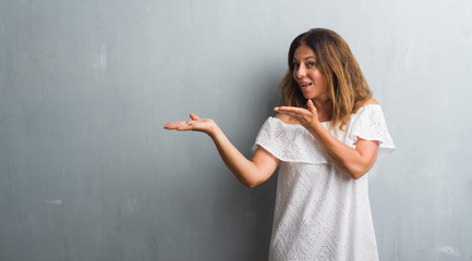 Middle age hispanic woman standing over grey grunge wall amazed and smiling to the camera while...