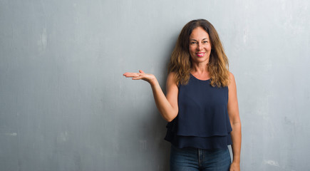 Middle age hispanic woman standing over grey grunge wall smiling cheerful presenting and pointing with palm of hand looking at the camera.
