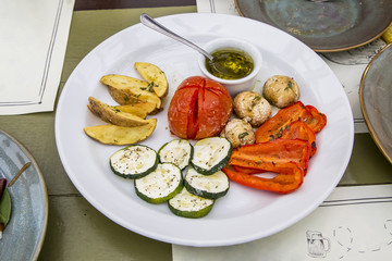 Assorted baked vegetables. Potatoes, cabbage, sweet peppers, tomatoes, champignons on a white plate with sauce. On the table