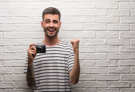 Young man holding vintage camera standing over white brick wall screaming proud and celebrating victory and success very excited, cheering emotion