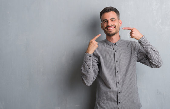 Young adult man standing over grey grunge wall smiling confident showing and pointing with fingers teeth and mouth. Health concept.