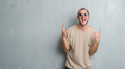 Young caucasian man over grey grunge wall wearing sunglasses shouting with crazy expression doing rock symbol with hands up. Music star. Heavy concept. - obrazy, fototapety, plakaty