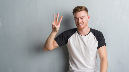 Young redhead man over grey grunge wall showing and pointing up with fingers number three while...