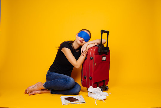 A Young Girl In A Black T-shirt Is Sleeping On A Big Red Suitcase, In Front Of Her Mask For Sleeping, Waiting For Her Plane