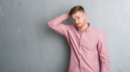 Young redhead man over grey grunge wall wearing pink shirt confuse and wonder about question. Uncertain with doubt, thinking with hand on head. Pensive concept.