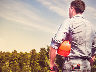 Worker with tools and an orange helmet standing against a background of green trees and blue sky....