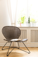 A rattan and metal chair with an open book on a seat standing on a wood flooring against a white wall and a sunny window in a living room interior. Real photo.