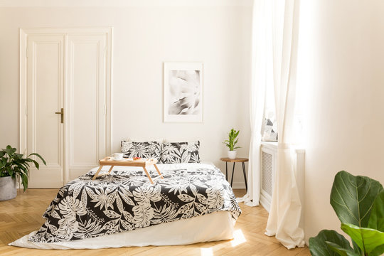 Comfortable big bed with white and black flower design bedding and a breakfast tray on in a wooden floor bright bedroom interior. Double door to another room in a background wall. Real photo.