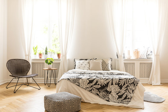 Comfy bedroom interior with a leaf motif bedding on a bed, a rattan chair and a black and white pouf standing on a herringbone parquet. Real photo.