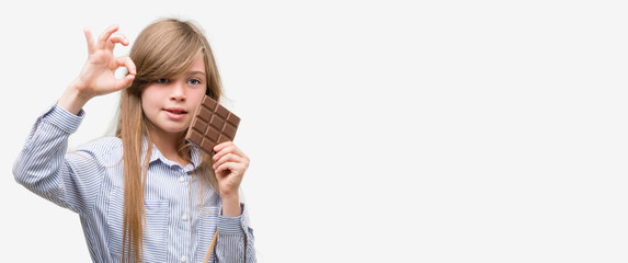Young blonde child holding chocolate bar doing ok sign with fingers, excellent symbol