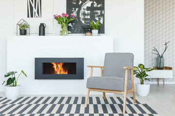 Wooden armchair on patterned carpet in modern flat interior with flowers above fireplace. Real photo