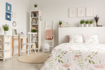 Real photo of a pastel bedroom interior with a double bed, floral sheets, dressing table, chair, shelf and plants