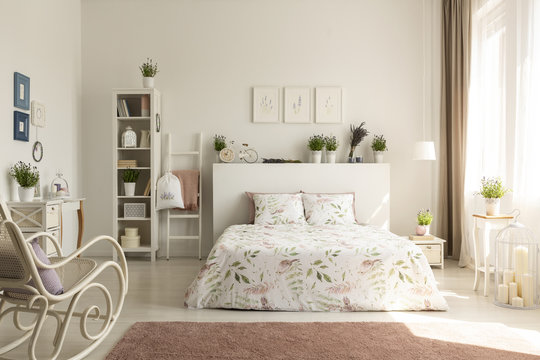 Real photo of a provencal bedroom interior with a double bed, floral sheets, shelves, plants and rocking chair