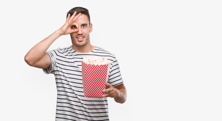 Handsome young man eating popcorn with happy face smiling doing ok sign with hand on eye looking...