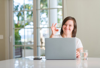 Down syndrome woman at home using computer laptop doing ok sign with fingers, excellent symbol