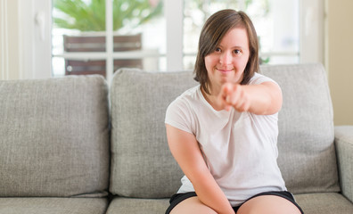 Down syndrome woman sitting on the sofa at home pointing with finger to the camera and to you, hand sign, positive and confident gesture from the front