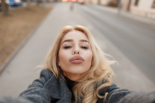 Fashionable young beautiful girl with big lips sends an air kiss and makes a photo of selfie on the street