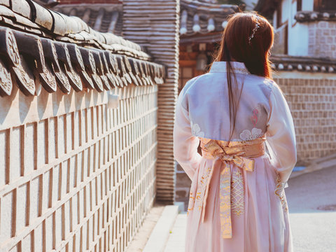 travel and tourist asia concept from backside long hair woman in korea traditional cloth (hanbok) relax, take picture in vintage town with rim light and soft focus background