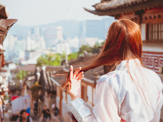 travel and tourist asia concept from backside of beauty long hair woman in korea traditional cloth (hanbok) stand, take picture and see to people in vintage town with soft focus background