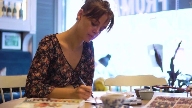 a young girl draws sketches on paper with a brush sitting at a table in a cafe. A fashion designer or tailor, animator, artist or Illustrator works in a cafe