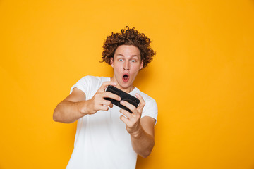 Young brunette guy in casual t-shirt playing online video games using mobile phone, isolated over...