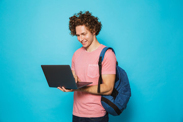 Image of attractive young guy with curly hair wearing casual clothing and backpack holding and using open laptop, isolated over blue background