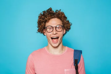 Fotobehang Image of geek guy with curly hair wearing glasses and backpack smiling at camera, isolated over blue background © Drobot Dean