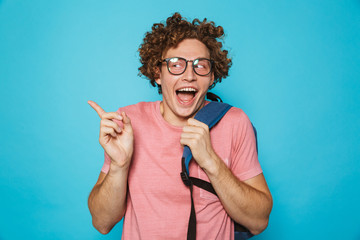 Photo of hipster geek man with curly hair wearing glasses and backpack smiling and gesturing aside, isolated over blue background